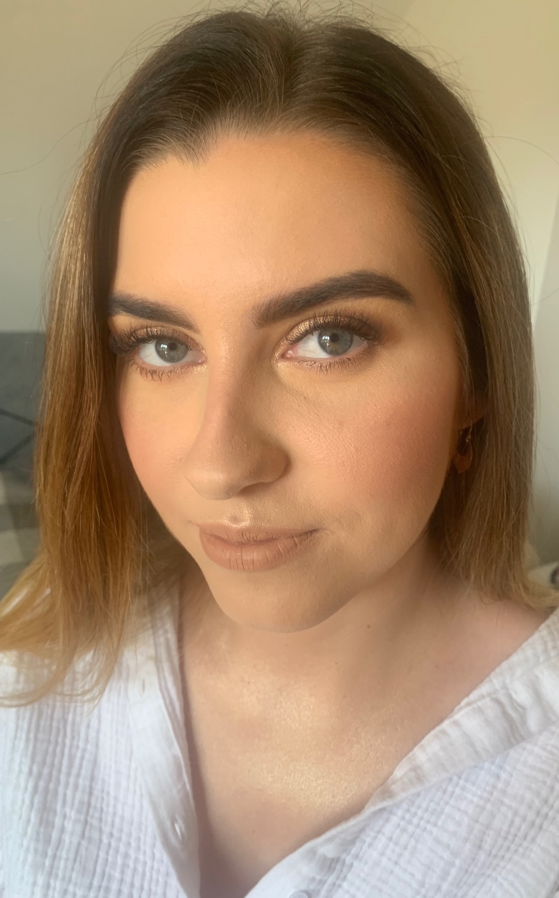 Portret kobiety z delikatnym makijażem, podkreślone brwi, złoty cień do powiek i matowa pomadka w odcieniu nude, widoczne kolczyki.