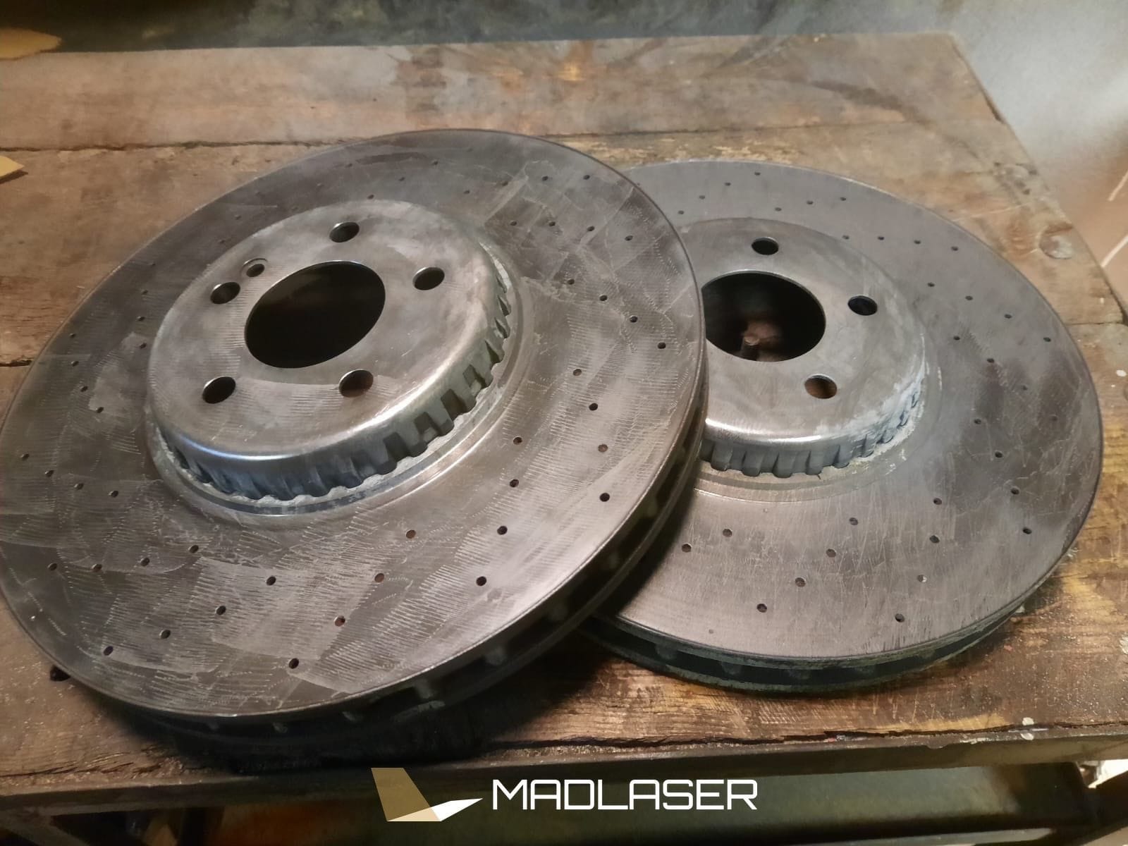 Dwa tarcze hamulcowe z nawierceniami, leżące na drewnianym stole po procesie obróbki, z widocznymi śladami po czyszczeniu i logo firmy w rogu zdjęcia.