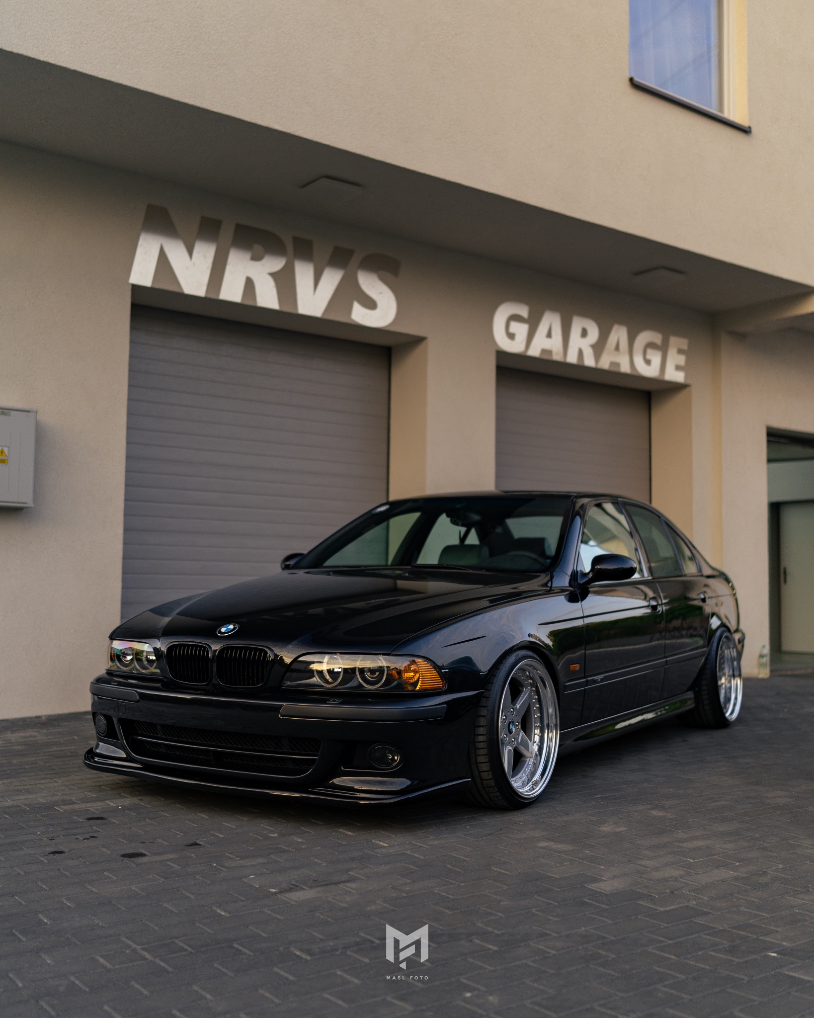 Czarna limuzyna BMW E39 540i zaparkowana przed garażem NRVS, z widocznymi srebrnymi felgami i obniżonym zawieszeniem.