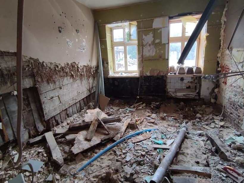 Wnętrze pomieszczenia w trakcie demontażu, widoczne odsłonięte fragmenty ścian z drewnianą konstrukcją, gruz na podłodze, okna z widokiem na zieleń na zewnątrz, fragmenty instalacji i elementy...