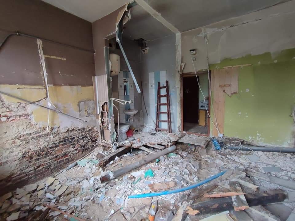 Wnętrze w trakcie gruntownego remontu: odsłonięta cegła, resztki płytek, gruz na podłodze, częściowo zdemontowana instalacja wodna w pomieszczeniu z toaletą, drabina oparta o ścianę.