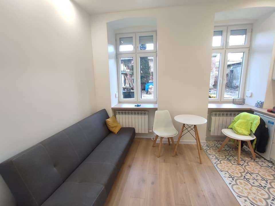 Jasne wnętrze kuchni w Zgierzu po remoncie: szara sofa z żółtą poduszką, biały stół z krzesłami, dwa okna z widokiem na podwórze, drewnopodobna podłoga i wzorzysty dywanik.