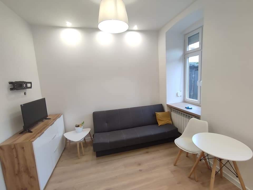 Jasne, nowocześnie urządzone wnętrze pokoju z szarą sofą, białym stolikiem kawowym i szafką RTV z drewnianym blatem, po remoncie.