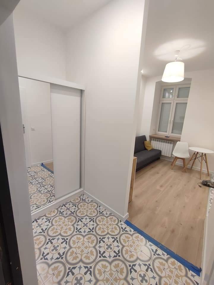 Wnętrze mieszkania po remoncie z nową, wzorzystą podłogą w przedpokoju, przechodzącą w drewnianą podłogę w salonie, białe ściany i przesuwne drzwi szafy wnękowej z lustrem.