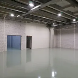 Wnętrze hali przemysłowej z jasnoszarą, jednolitą posadzką, widoczne kolumny, ściany z białej cegły i instalacje pod sufitem.