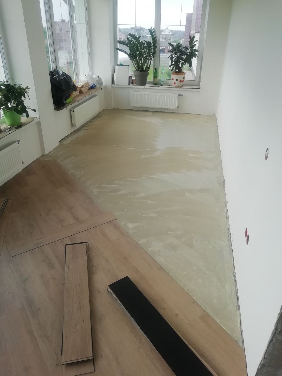 Proces układania paneli podłogowych w jasnym pokoju z dużymi oknami, widoczna część podłogi pokryta wylewką, a część z ułożonymi panelami i kilkoma panelami przygotowanymi do montażu.