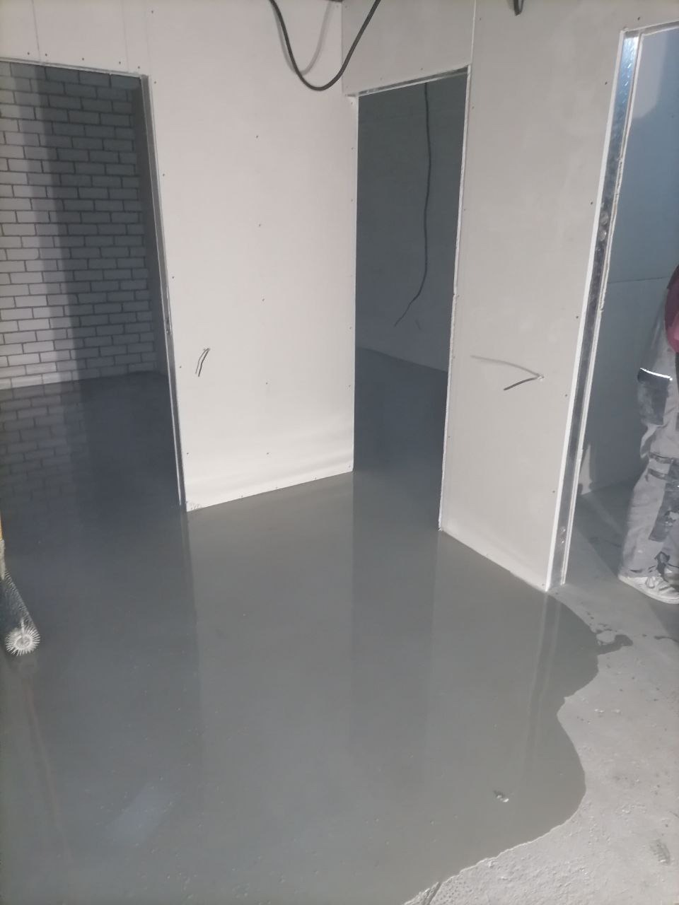 Świeżo wylana, szara posadzka samopoziomująca w pomieszczeniu w Kobyłce, z widocznym wałkiem do odpowietrzania i fragmentem pracownika w tle, przygotowana do dalszych prac wykończeniowych.