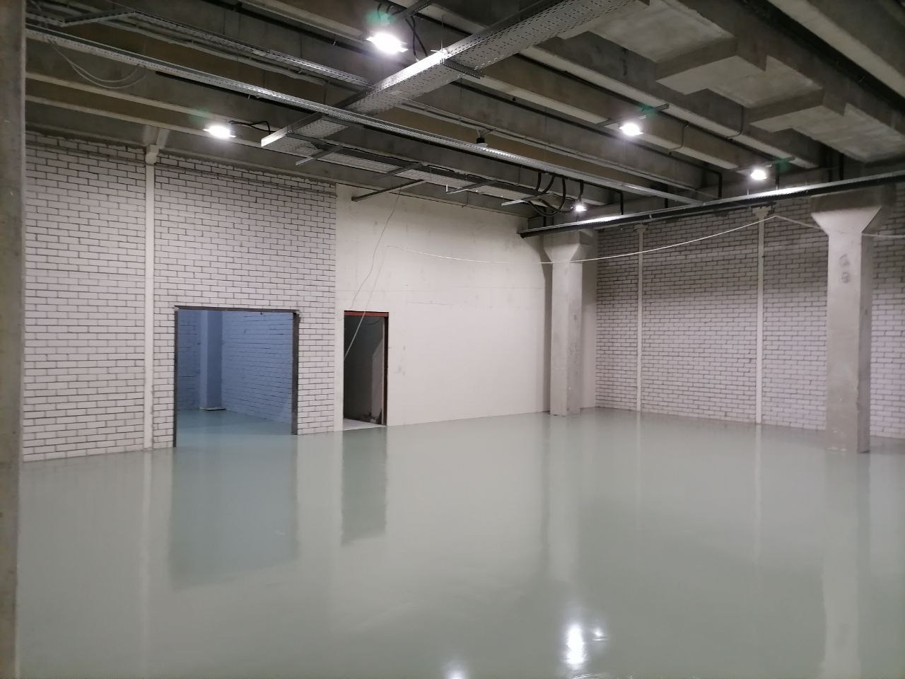 Wnętrze hali przemysłowej z jasnoszarą, jednolitą posadzką, widoczne kolumny, ściany z białej cegły i instalacje pod sufitem.