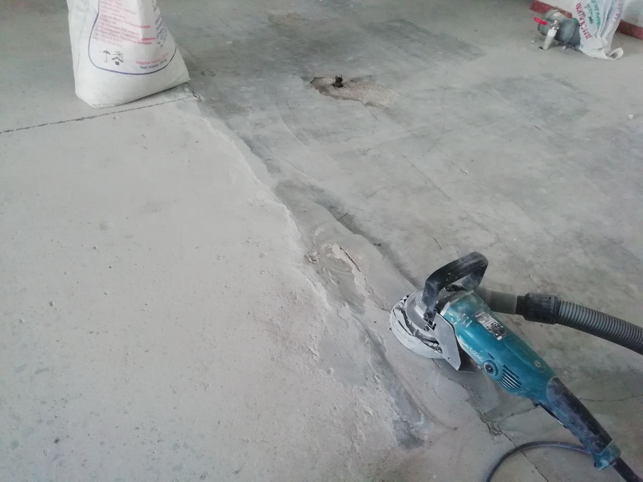 Szlifowanie nierówności posadzki jastrychowej szlifierką kątową z odkurzaczem, widoczny worek z zaprawą i ślady po szlifowaniu.