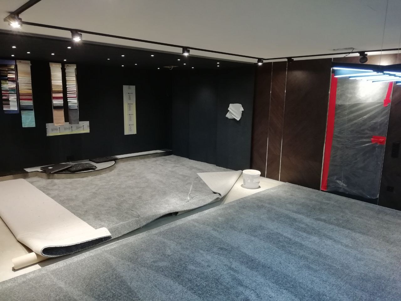 Przygotowanie szarej podłogi w pomieszczeniu z czarnymi ścianami i oświetleniem punktowym, widoczny częściowo zwinięty biały materiał oraz wiadro z farbą.