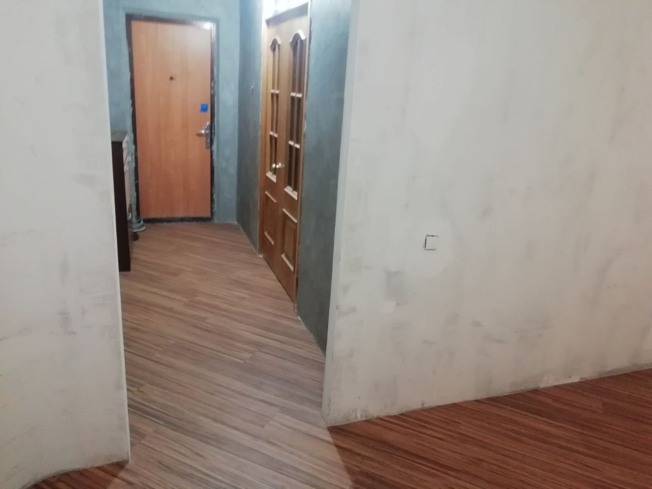 Korytarz z nową podłogą imitującą drewno, ułożoną pod kątem. Widoczne drzwi wejściowe i wewnętrzne. Ściany w trakcie wykańczania, z widocznymi śladami po pracach remontowych.