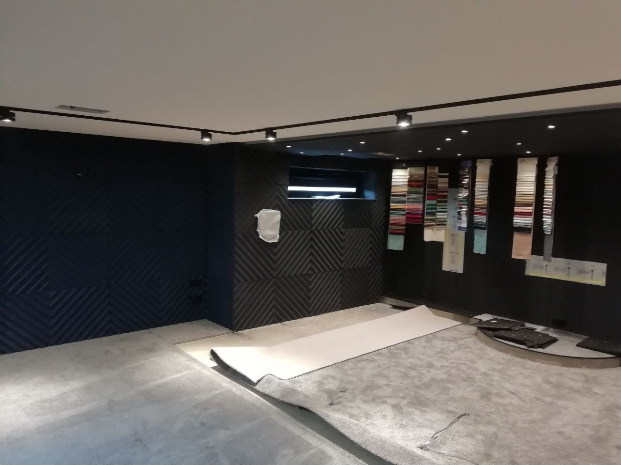 Wnętrze w trakcie prac wykończeniowych, z szarą posadzką wylewaną, częściowo przykrytą ochronną folią, obok próbki kolorów na ścianie i dekoracyjne panele ścienne w geometryczny wzór.