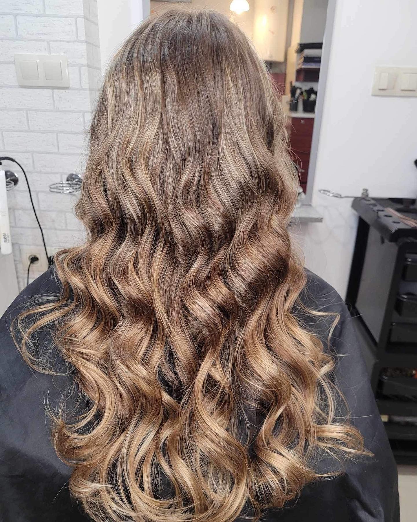 Długie, falowane włosy z delikatnym ombre w odcieniach blondu i brązu, widoczne od tyłu na tle ściany z białej cegły i czarnego wózka fryzjerskiego.