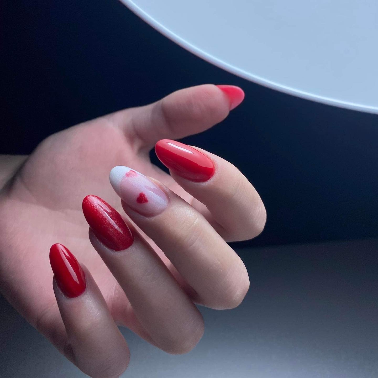 Dłoń z czerwonym manicure hybrydowym, jeden paznokieć z białym ombre i czerwonym serduszkiem, owalny kształt paznokci, oświetlenie lampą UV.