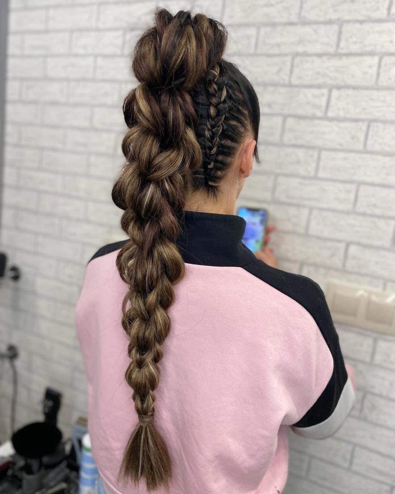 Widok z tyłu na kobiecą fryzurę z ciemnych włosów z blond pasemkami, upiętą w wysoki kok i zaplecioną w gruby warkocz typu dobierany, z dwoma mniejszymi warkoczami po bokach głowy, na tle białej...