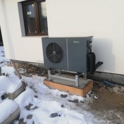 Arlok - Szara pompa ciepła zamontowana na metalowej konstrukcji przy białej ścianie budynku z brązowym oknem, widoczny śnieg na ziemi.