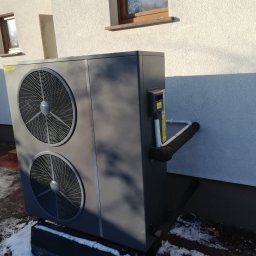 Arlok - Szara pompa ciepła typu powietrze-woda zainstalowana na zewnątrz budynku z elewacją w jasnym kolorze. Widoczne dwa wentylatory, rury izolowane termicznie oraz fragment ośnieżonego terenu.