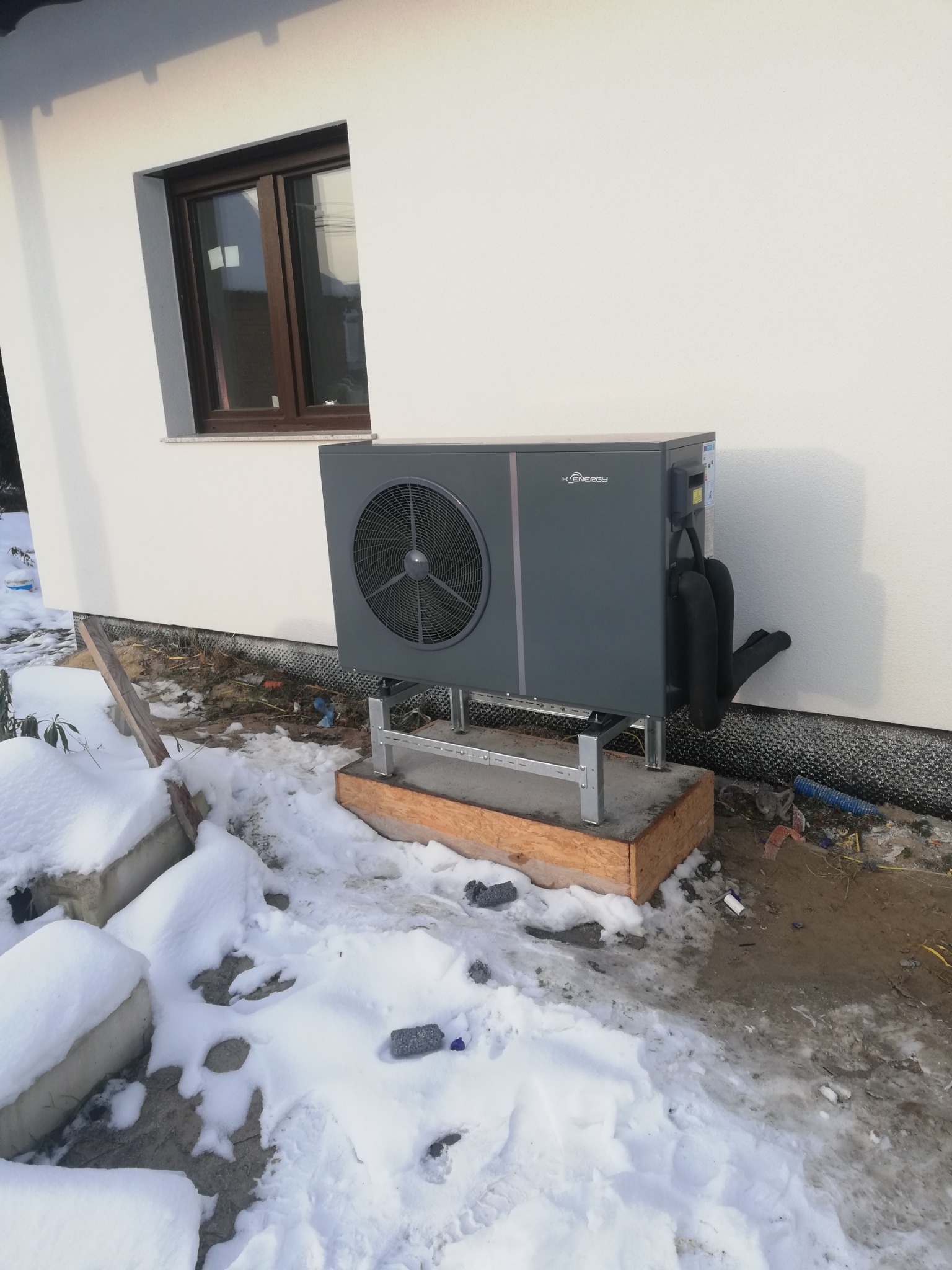 Szara pompa ciepła zamontowana na metalowej konstrukcji przy białej ścianie budynku z brązowym oknem, widoczny śnieg na ziemi.