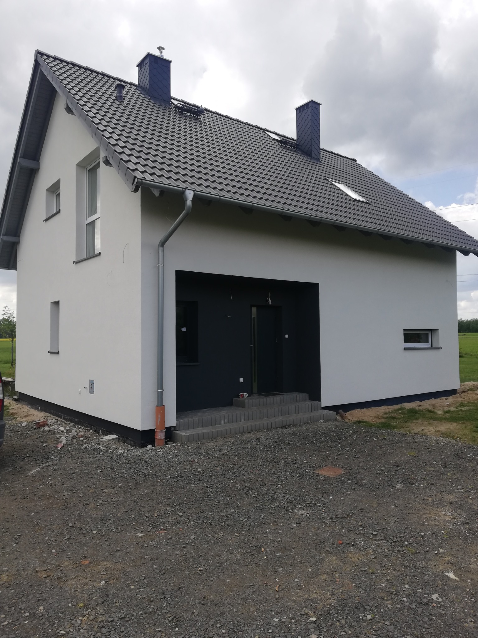 Nowoczesny, jednorodzinny dom z jasną elewacją, ciemną stolarką okienną i grafitowym dachem, widoczny z zewnątrz na tle pochmurnego nieba i zielonego krajobrazu.