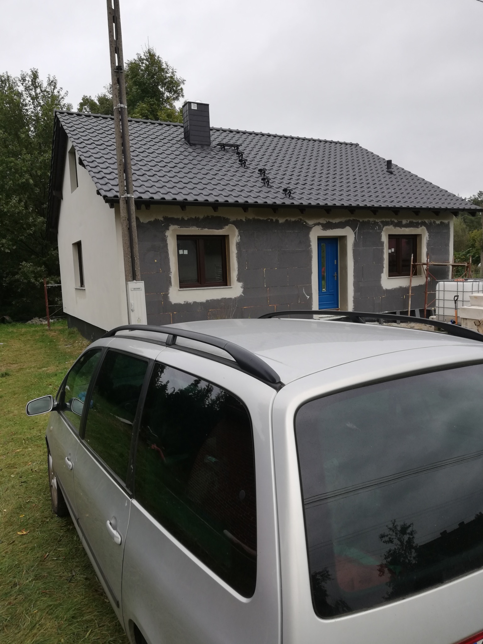 Dom jednorodzinny w trakcie prac elewacyjnych, częściowo ocieplony, z niebieskimi drzwiami wejściowymi, widziany zza srebrnego samochodu kombi.