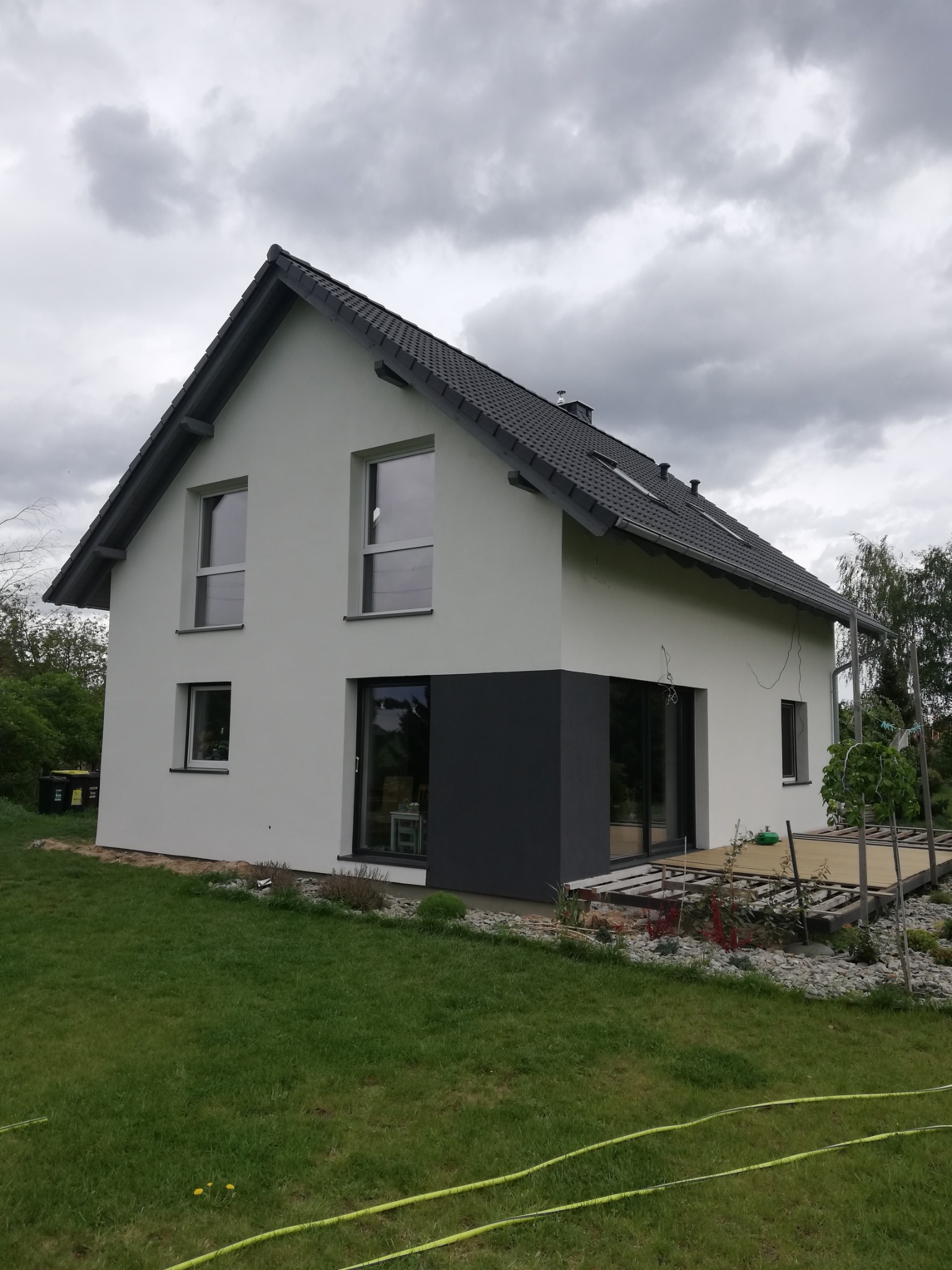 Dwukondygnacyjny dom z ocieploną białą elewacją, szarą podmurówką i ciemną dachówką, widok z trawnika z żółtym wężem ogrodowym, częściowo wykończony taras z desek.