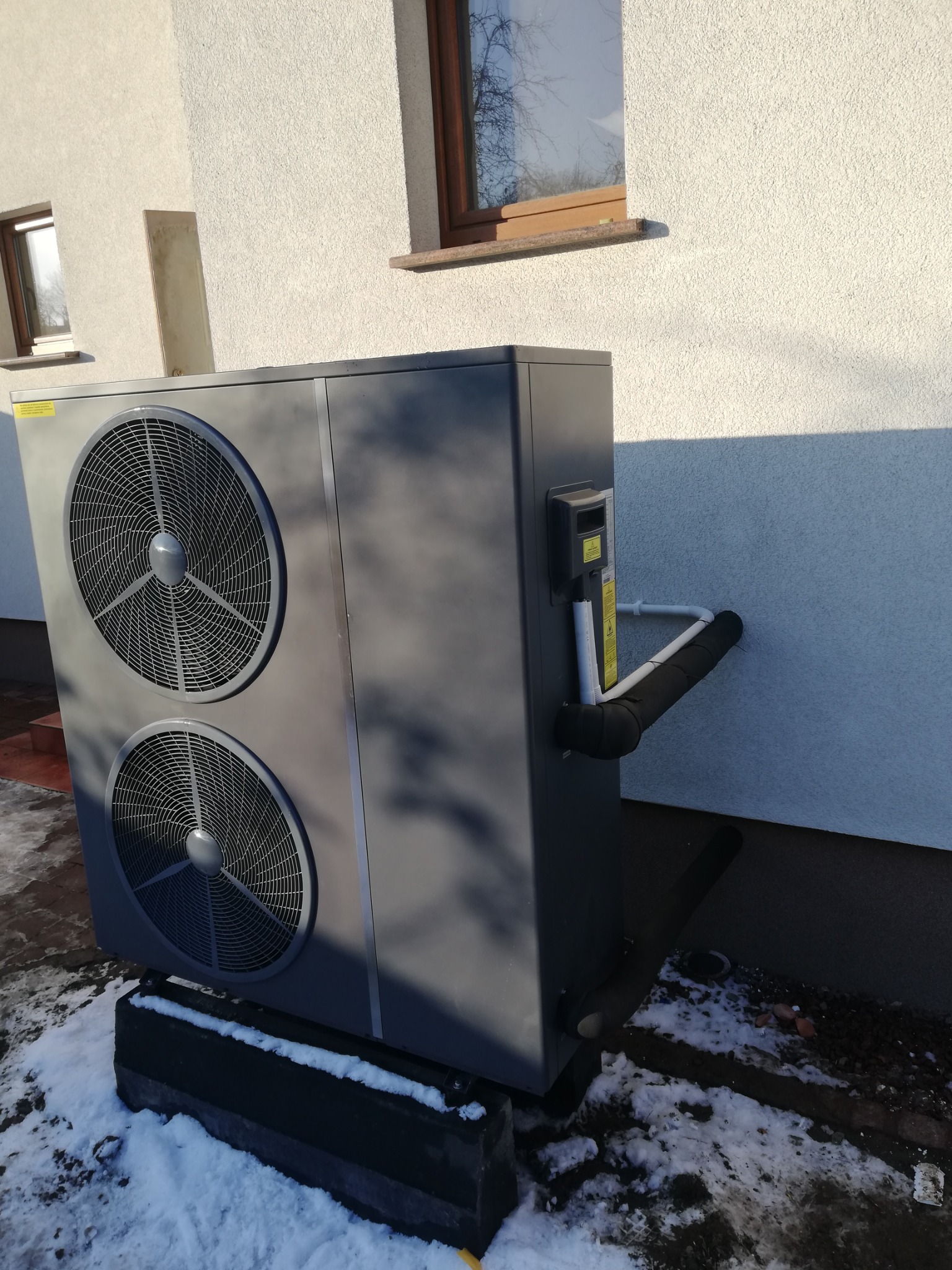 Szara pompa ciepła typu powietrze-woda zainstalowana na zewnątrz budynku z elewacją w jasnym kolorze. Widoczne dwa wentylatory, rury izolowane termicznie oraz fragment ośnieżonego terenu.