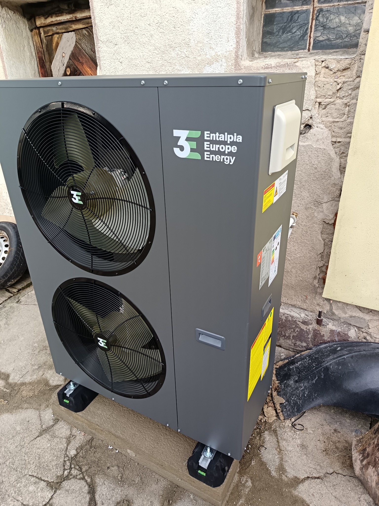 Szara pompa ciepła marki Entalpia Europe Energy, ustawiona na czarnych podkładkach z poziomicą, z widocznymi dwoma wentylatorami, na zewnątrz budynku z elementami muru i okna w tle.