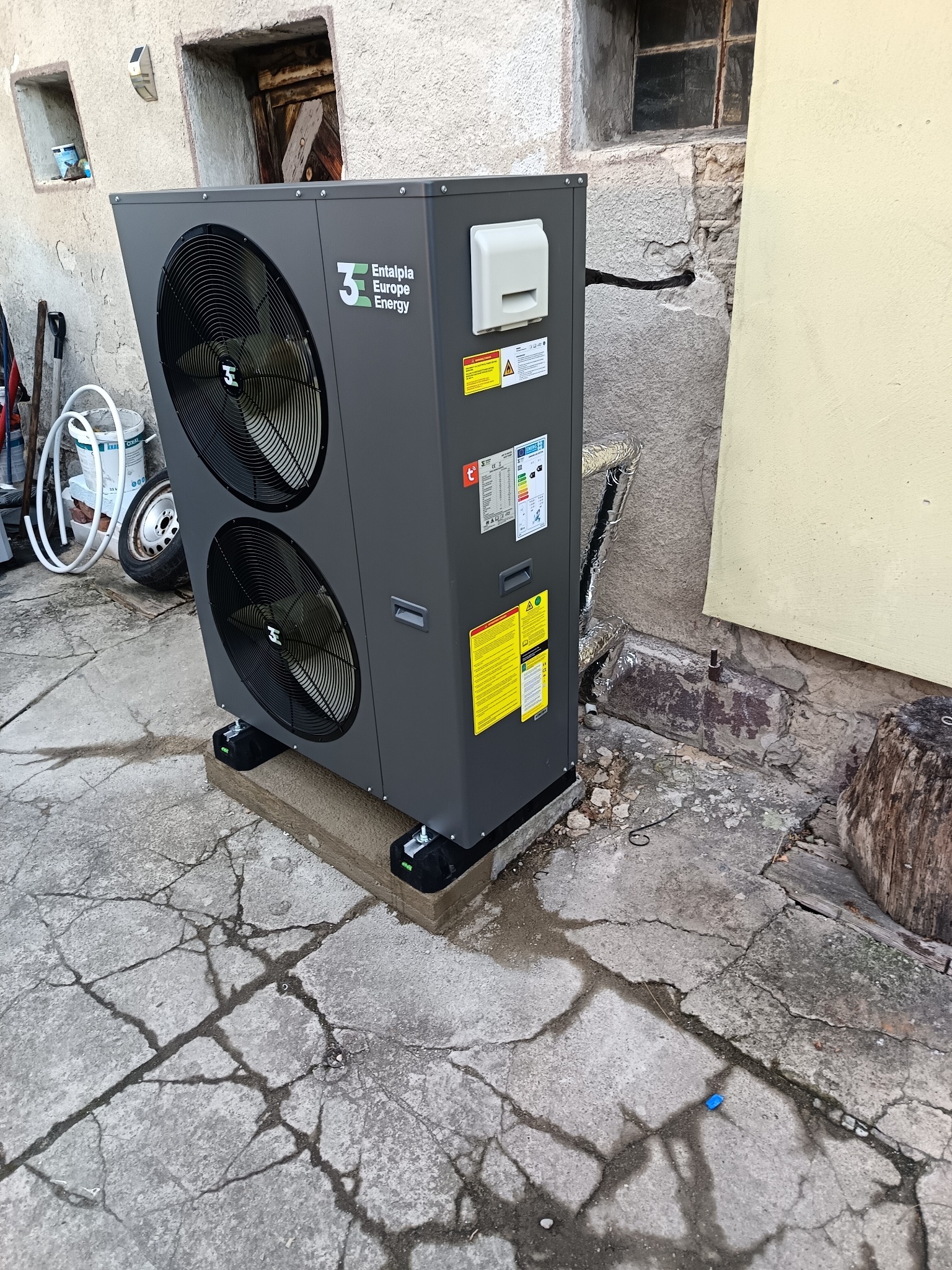 Szara pompa ciepła z dwoma wentylatorami marki Entalpia Europe Energy, zamontowana na betonowej podstawie przy szarej, starej ścianie z małymi oknami.
