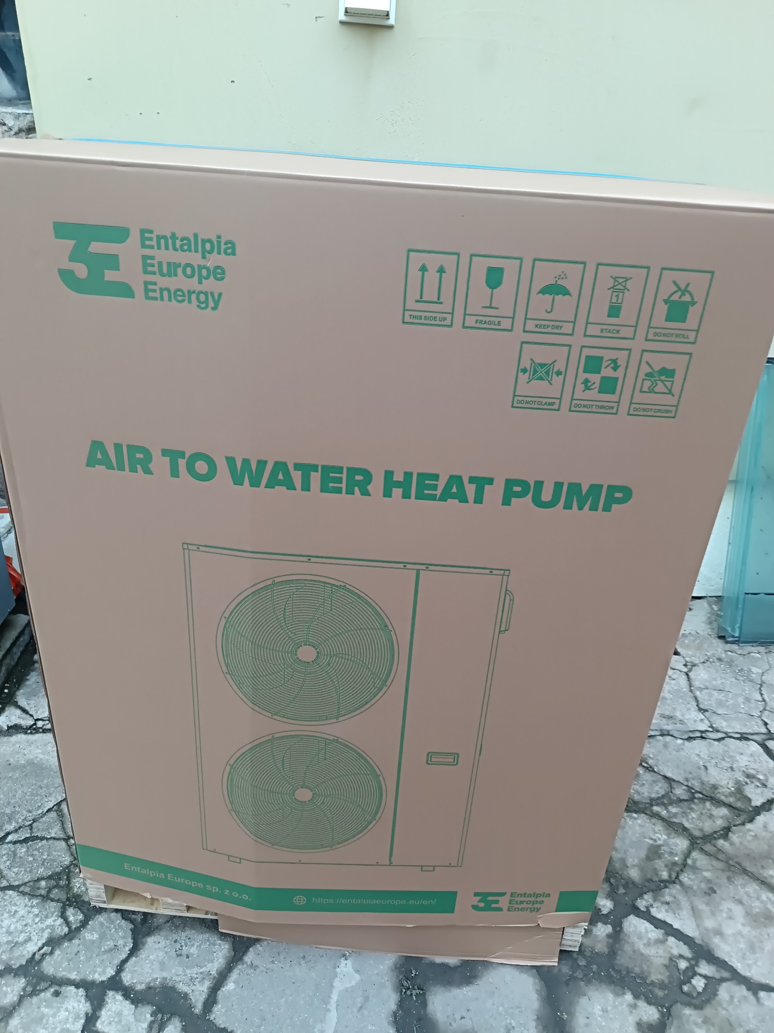 Kartonowe opakowanie pompy ciepła powietrze-woda, z widocznym schematycznym rysunkiem urządzenia na boku, umieszczone na palecie.