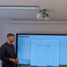 Prelegent prezentuje wyniki badań na interaktywnej tablicy podczas wykładu. Na ekranie widoczna tabela z danymi statystycznymi. Na suficie zamontowany projektor.