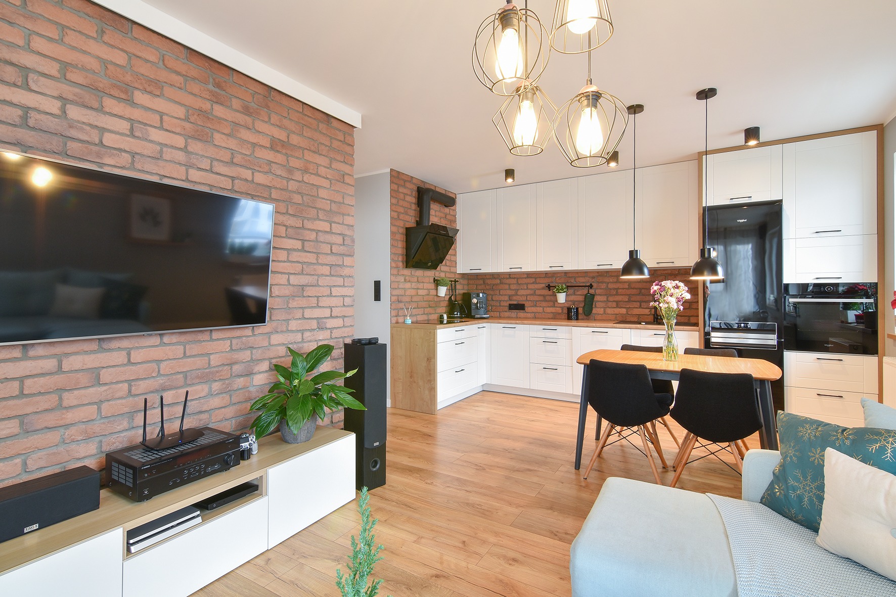 Nowoczesne wnętrze: ceglana ściana z telewizorem, białe meble kuchenne, drewniany parkiet, stół z krzesłami i dekoracyjne lampy wiszące