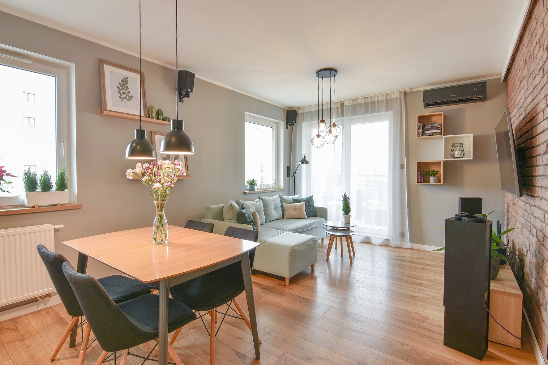 Jasne, przestronne wnętrze mieszkania z drewnianą podłogą, szarą sofą, stołem z kwiatami i ceglaną ścianą z telewizorem. Widoczne okna z roślinami, nowoczesne oświetlenie i elementy dekoracyjne...