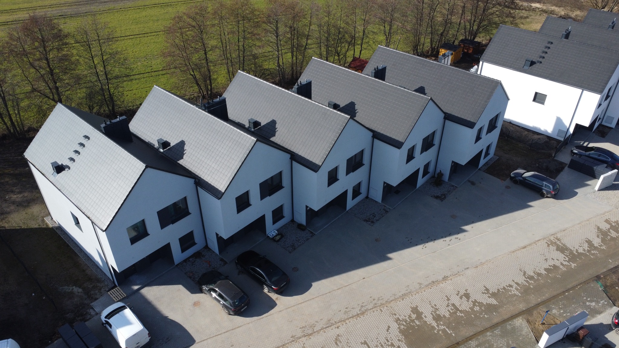 Nowoczesne szeregówki z szarymi dachami i białymi elewacjami, widok z lotu ptaka, zaparkowane samochody, surowy krajobraz.