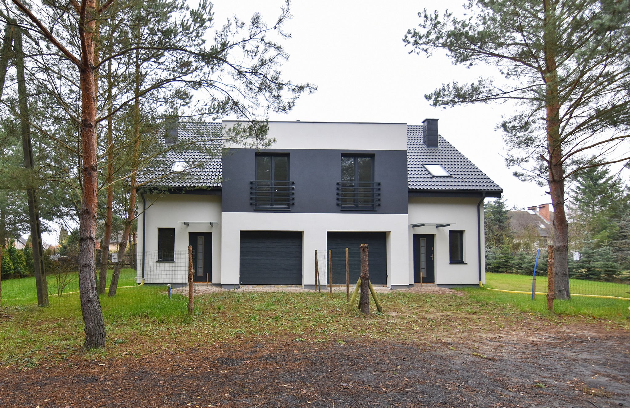 Dwukondygnacyjny dom bliźniak z szarą dachówką i podwójnym garażem, otoczony drzewami iglastymi i liściastymi, widok z zewnątrz na tle pochmurnego nieba.