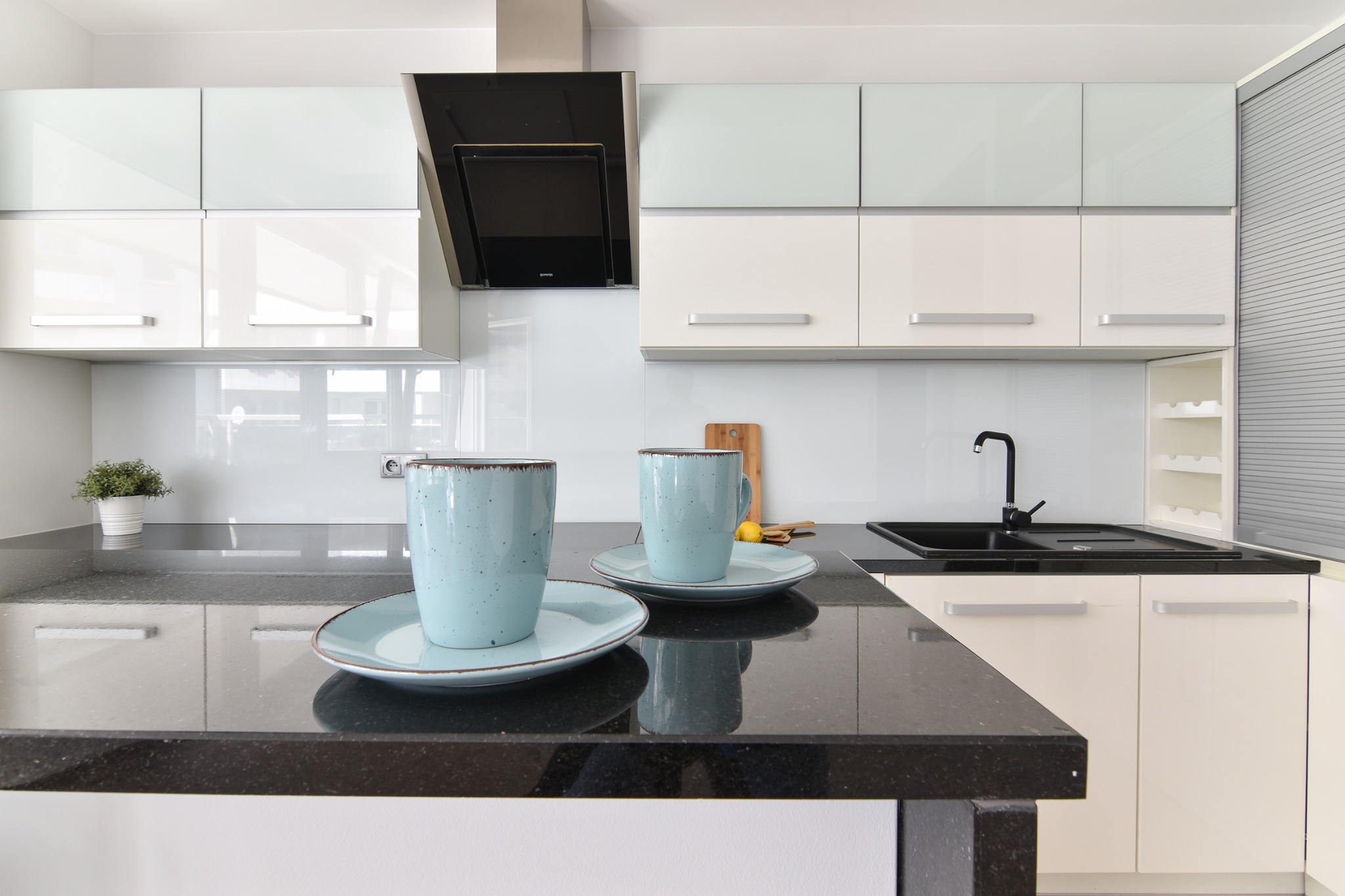 Nowoczesna kuchnia z białymi szafkami, czarnym blatem i dwiema turkusowymi filiżankami na pierwszym planie.