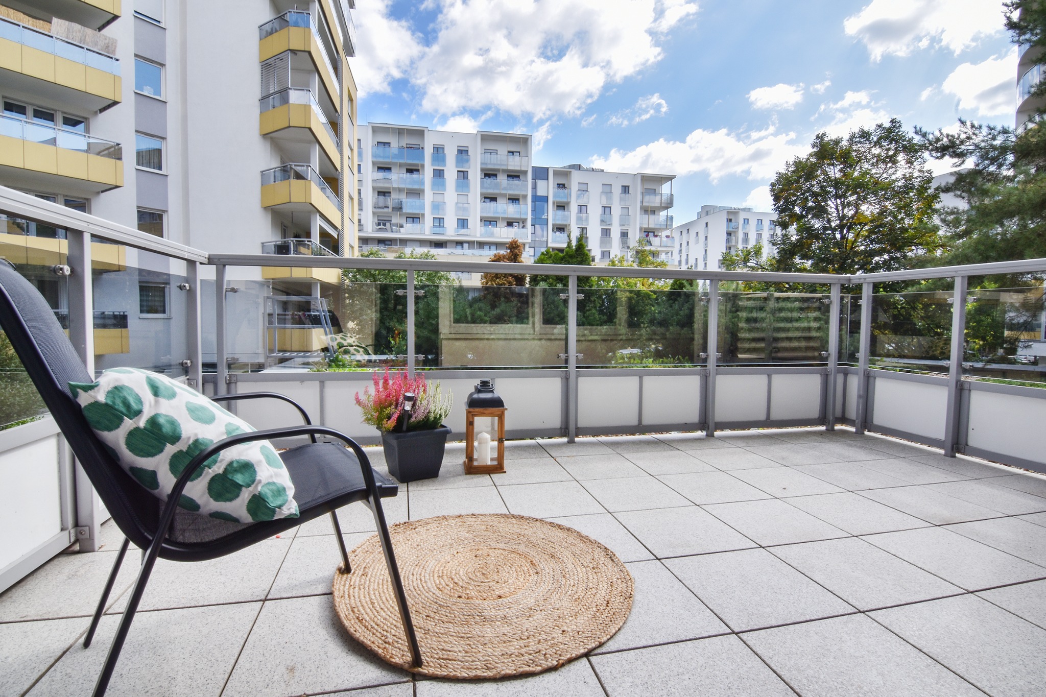 Umeblowany balkon z krzesłem z poduszką w zielone grochy, okrągłym jutowym dywanikiem, lampionem ze świecą i doniczką z wrzosem, na tle budynków mieszkalnych i błękitnego nieba.