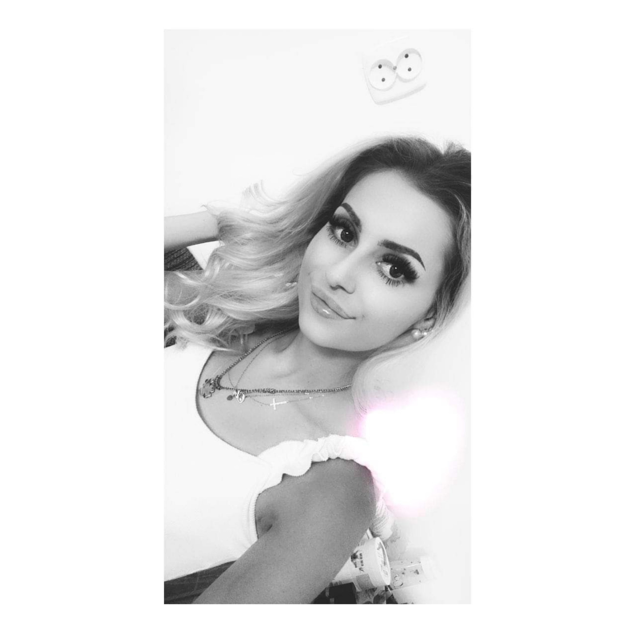 Portret kobiety z blond włosami i wyrazistym makijażem, leżącej na białym tle z widocznymi gniazdkami elektrycznymi w tle i kosmetykami obok.