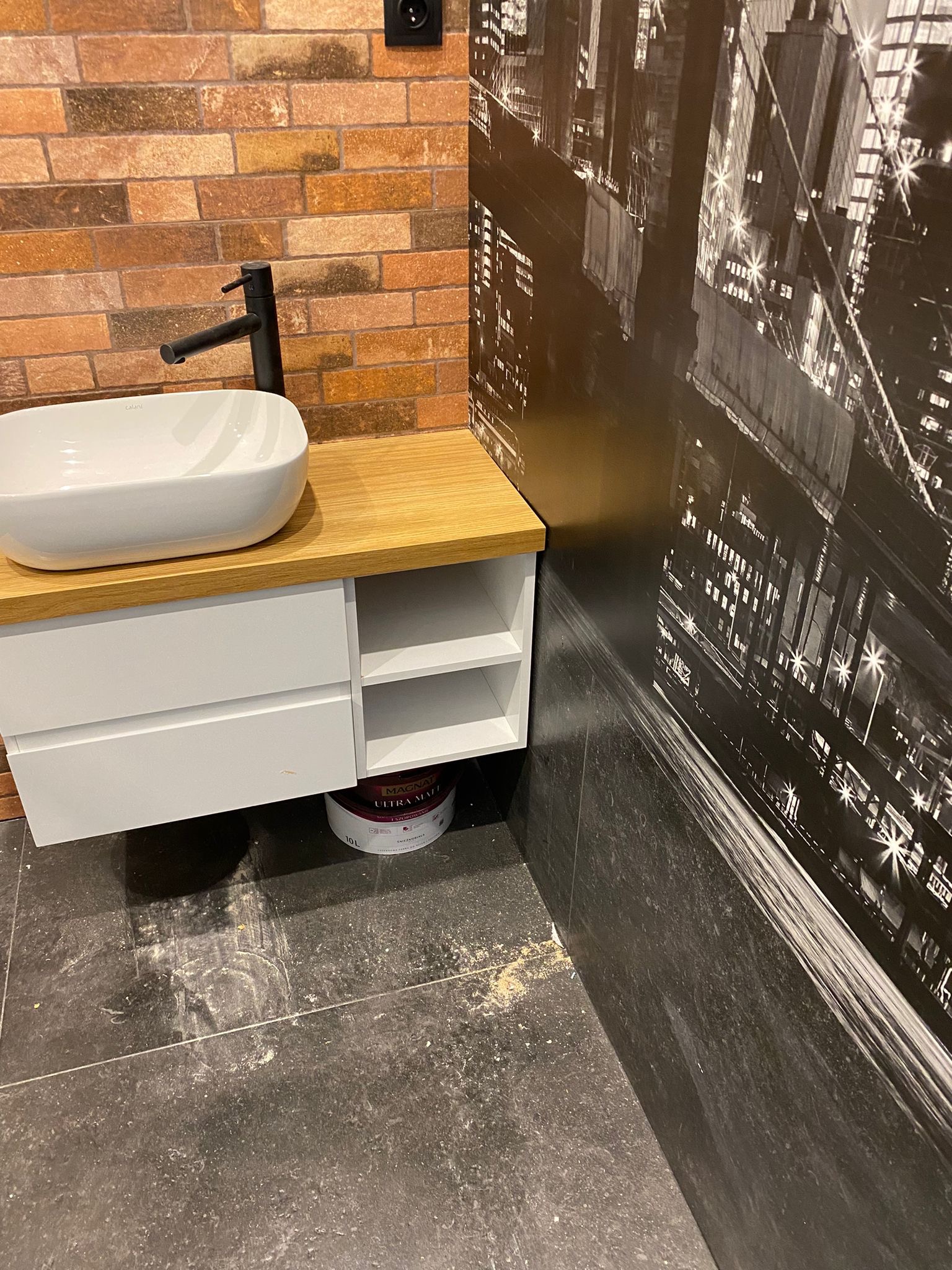 Nowoczesna łazienka z umywalką nablatową na szafce z drewnianym blatem, ceglaną ścianą i fototapetą z panoramą miasta, widoczny pojemnik z farbą pod szafką i ślady po remoncie na podłodze.