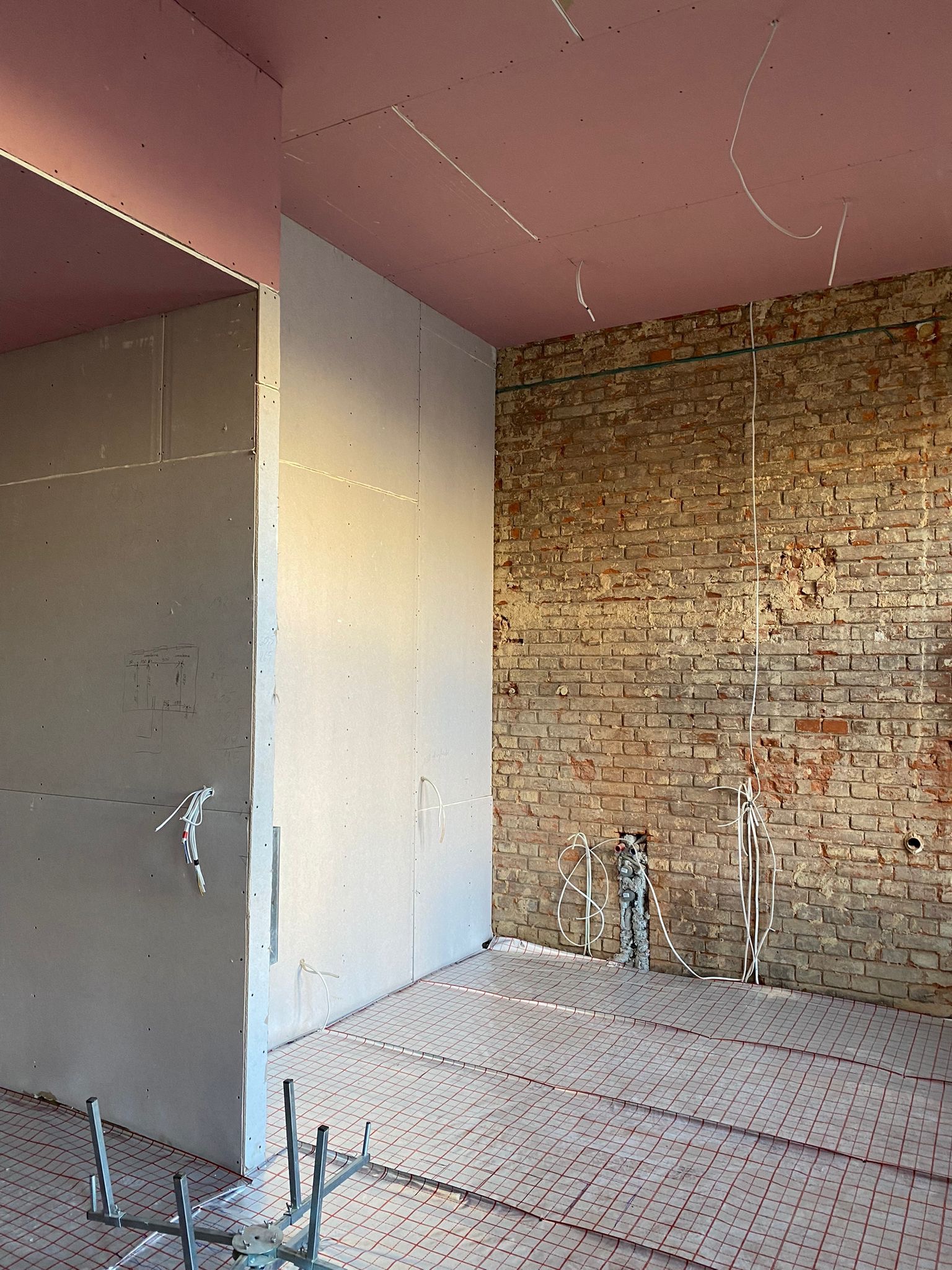 Wnętrze w trakcie prac w Bytomiu: surowa ceglana ściana kontrastuje z płytami gipsowo-kartonowymi na suficie i ścianach, widoczne instalacje elektryczne i ogrzewanie podłogowe.