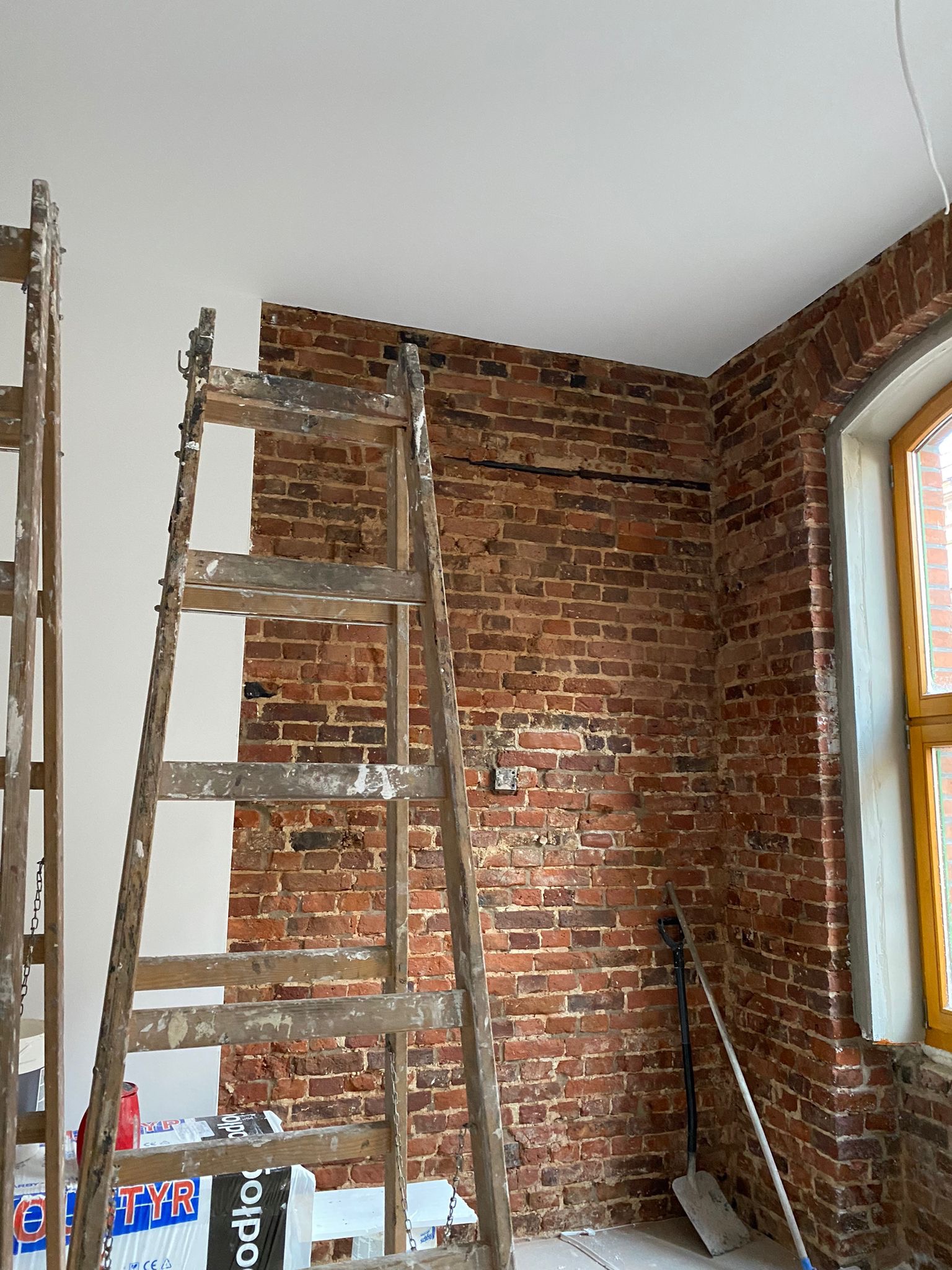 Częściowo pomalowana ceglana ściana z drewnianą drabiną, widoczne ślady farby, szpachelka i łopata oparta o ścianę, okno z drewnianą ramą.