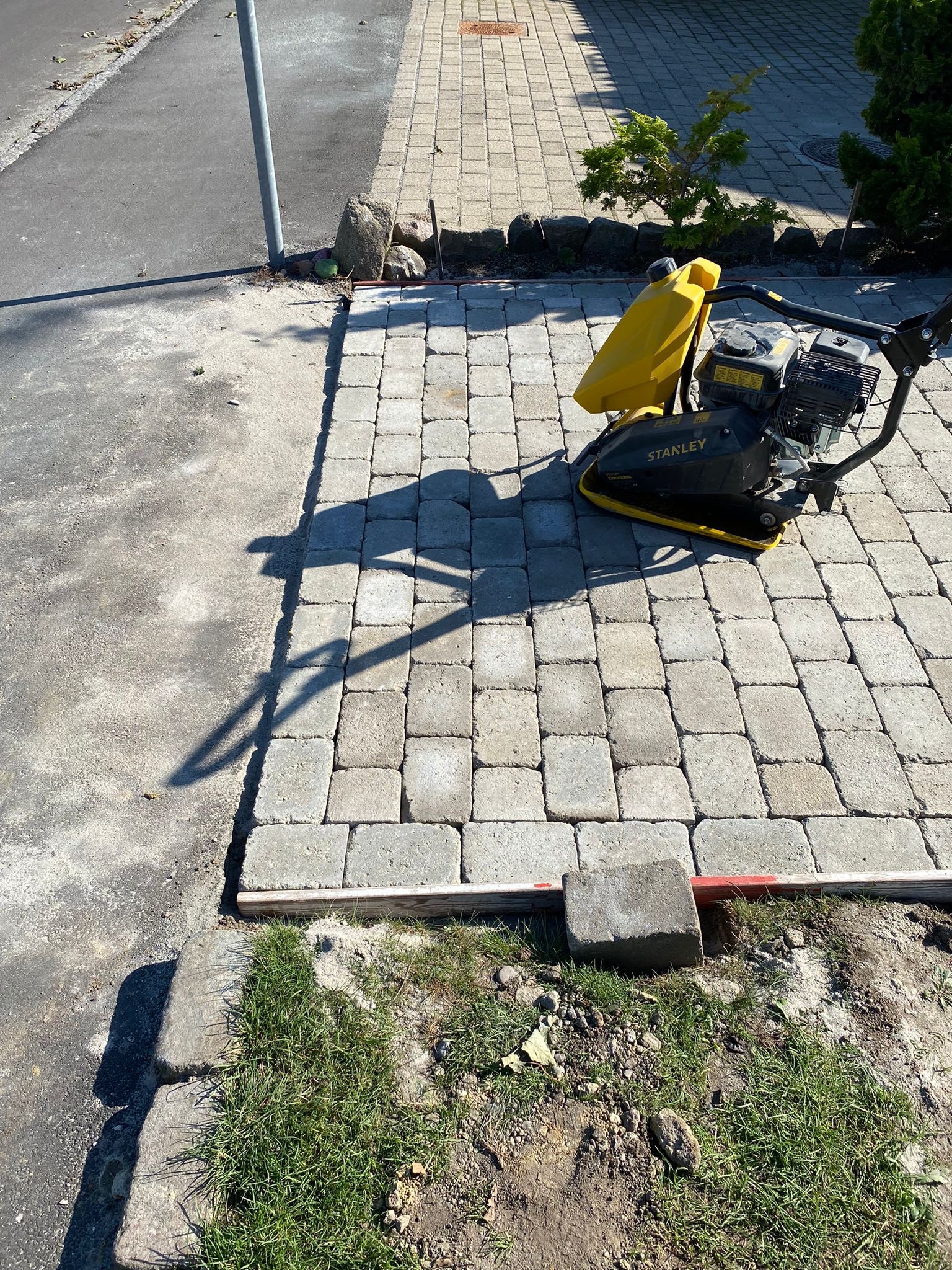 Żółto-czarna zagęszczarka Stanley na szarej kostce brukowej, obok trawnik i betonowa nawierzchnia, widoczne drewniane ograniczniki.