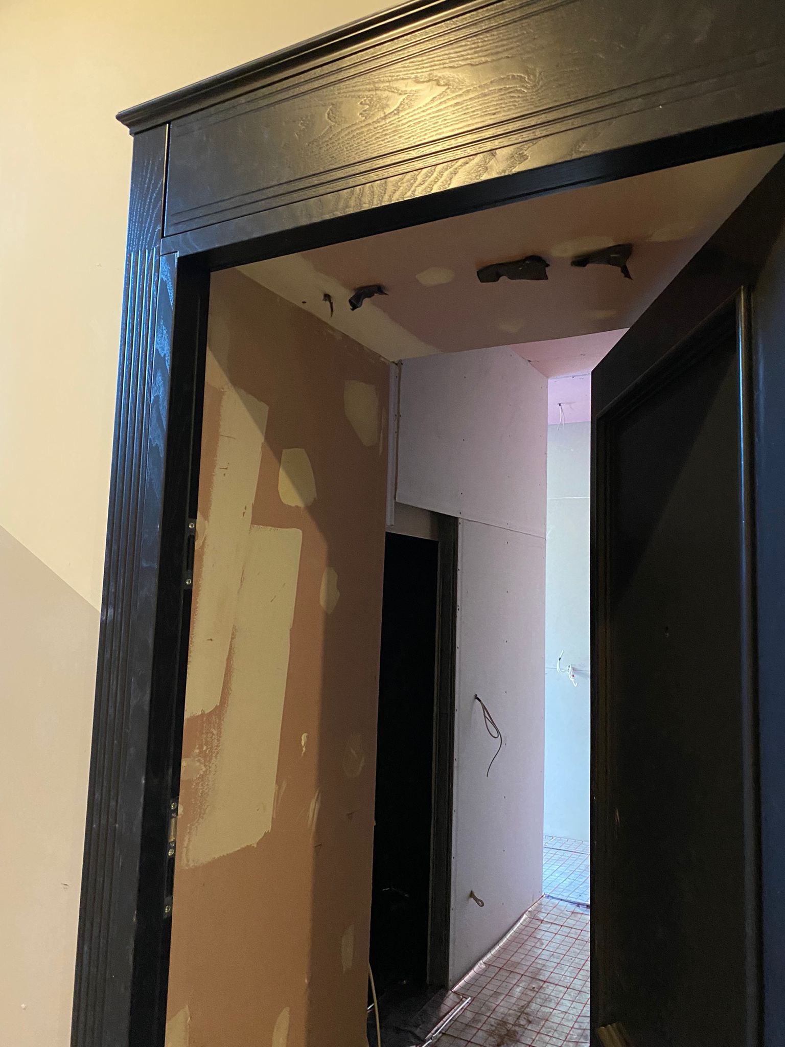 Wnętrze w trakcie prac wykończeniowych: widok przez futrynę z ciemnego drewna na ścianę z częściowo nałożonymi łatami gipsowymi, sufit z widocznymi mocowaniami oświetlenia i podłogę z instalacją...