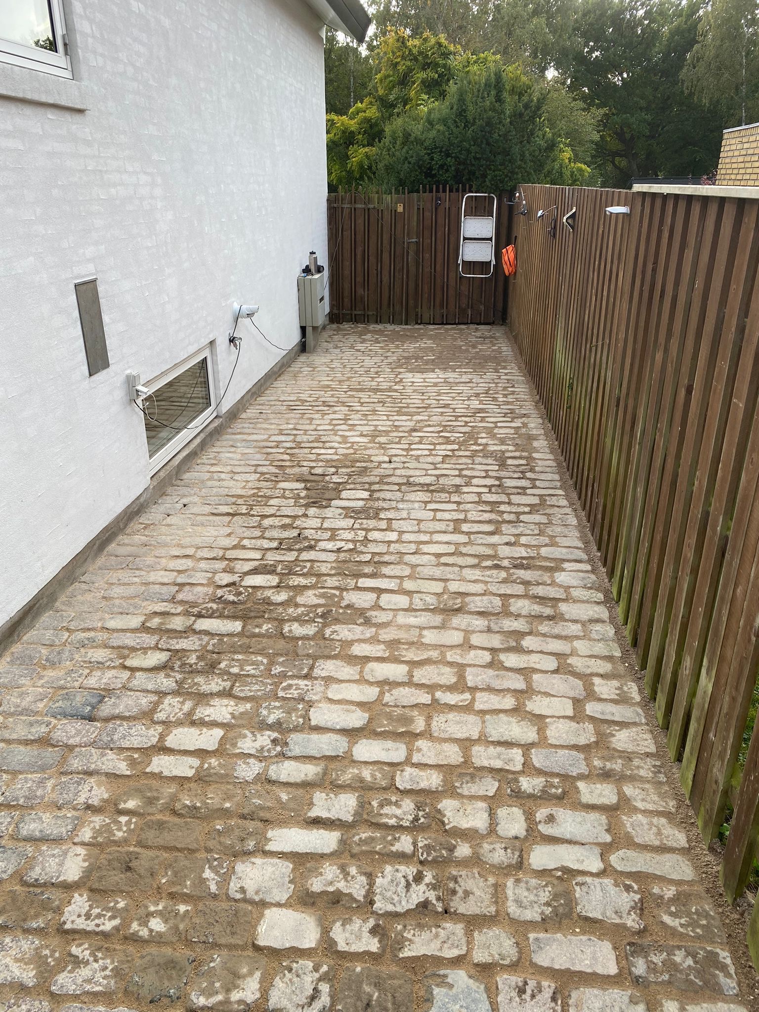 Ułożona z kostki brukowej ścieżka prowadząca wzdłuż białego budynku i drewnianego płotu z drabinką, widoczna instalacja elektryczna na ścianie.