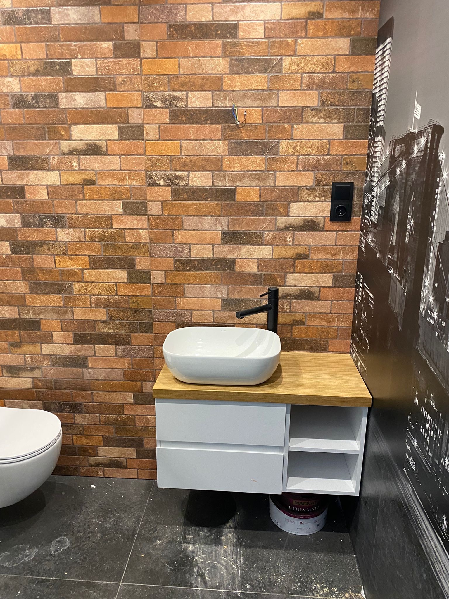 Nowoczesna łazienka z umywalką nablatową na szafce z drewnianym blatem, ściana wyłożona ceglastą płytką, obok toaleta i tapeta z motywem miasta, widoczna puszka farby.
