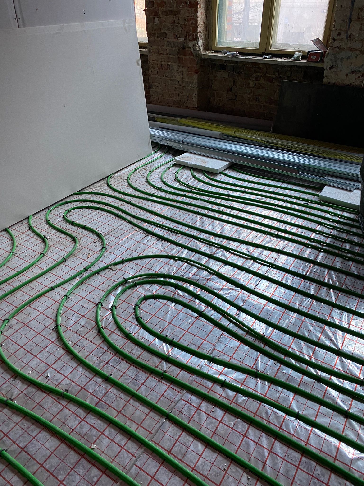 Układanie zielonych rur ogrzewania podłogowego na folii z czerwoną kratką w surowym stanie, widoczny fragment ściany z cegły i okno w tle.