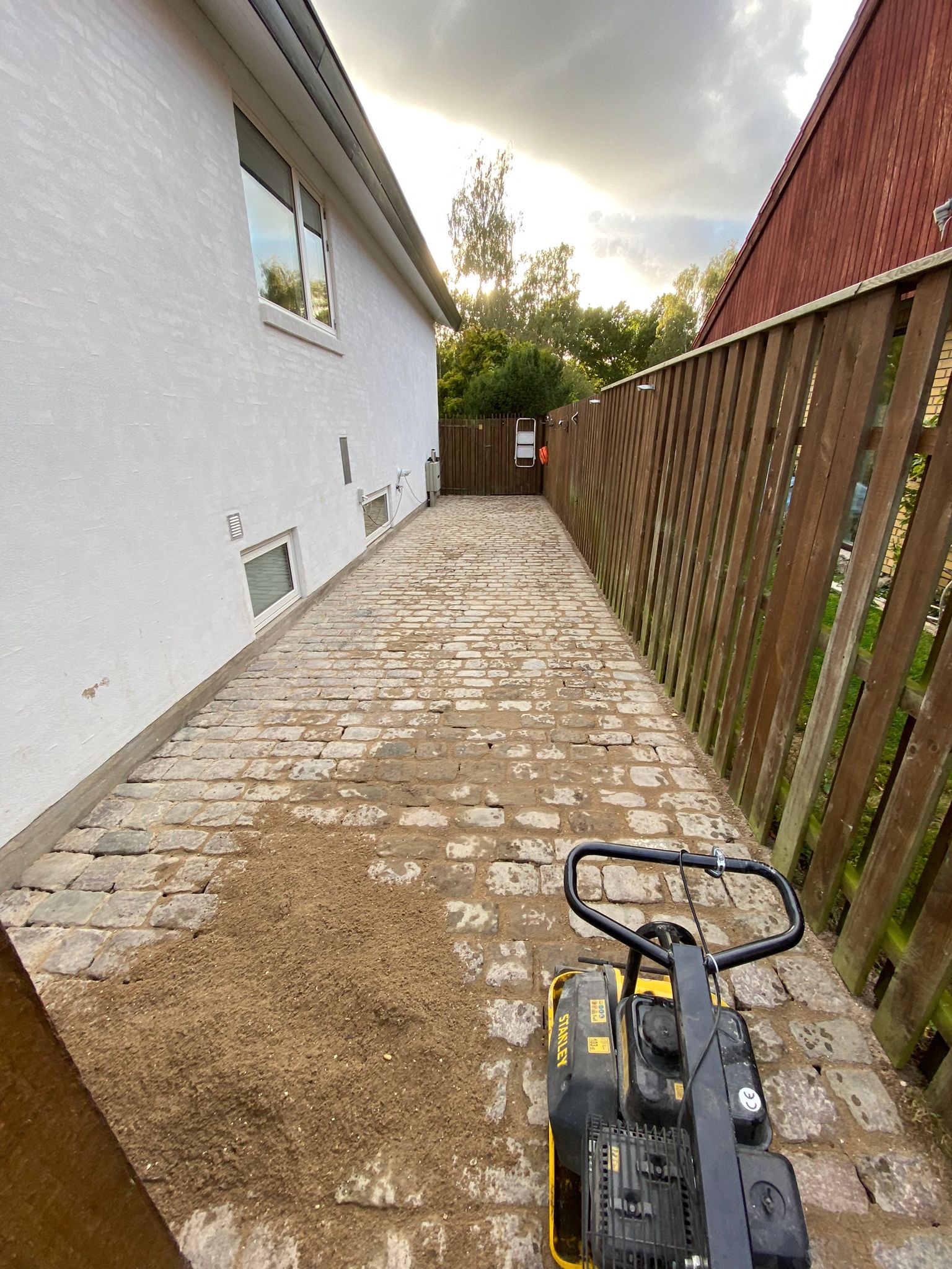 Układanie kostki brukowej na podwórku, ubijarka Stanley na pierwszym planie, widoczny fragment białego budynku i drewnianego ogrodzenia.
