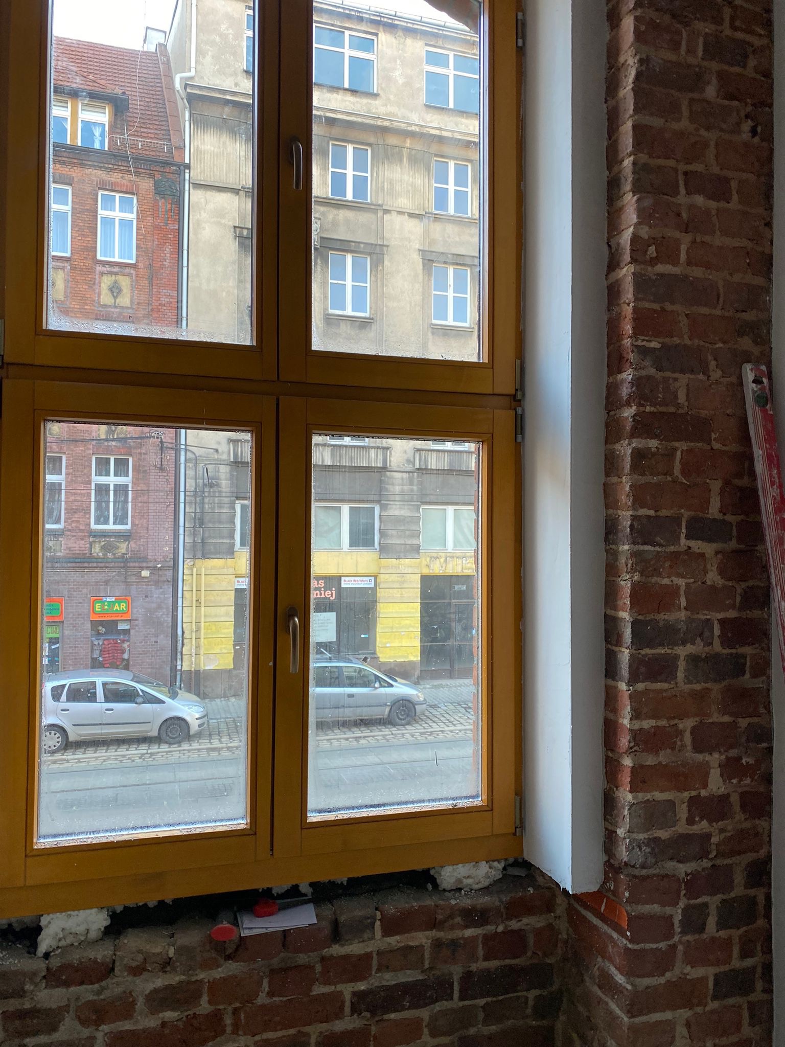 Żółte okno z widokiem na ulicę w trakcie prac wykończeniowych, widoczna cegła i pianka montażowa.