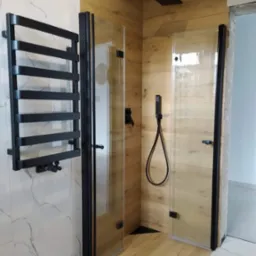 Nowoczesna kabina prysznicowa z czarnymi elementami, drewnopodobne panele ścienne, grzejnik drabinkowy w kolorze czarnym na marmurowej ścianie.