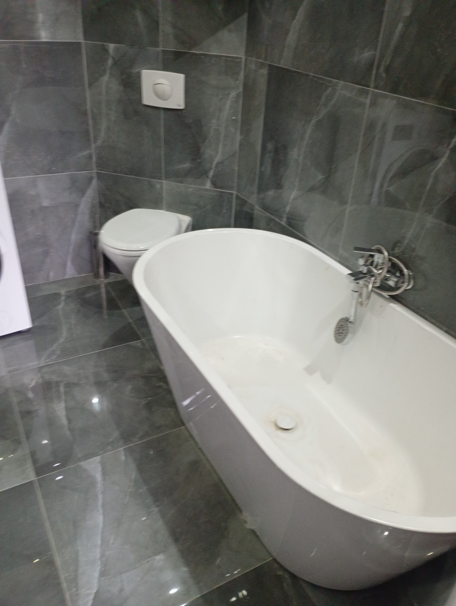 Nowoczesna łazienka z wolnostojącą wanną, szarą marmurową okładziną ścian i podłogi oraz minimalistyczną armaturą. Widoczny fragment pralki i toaleta.