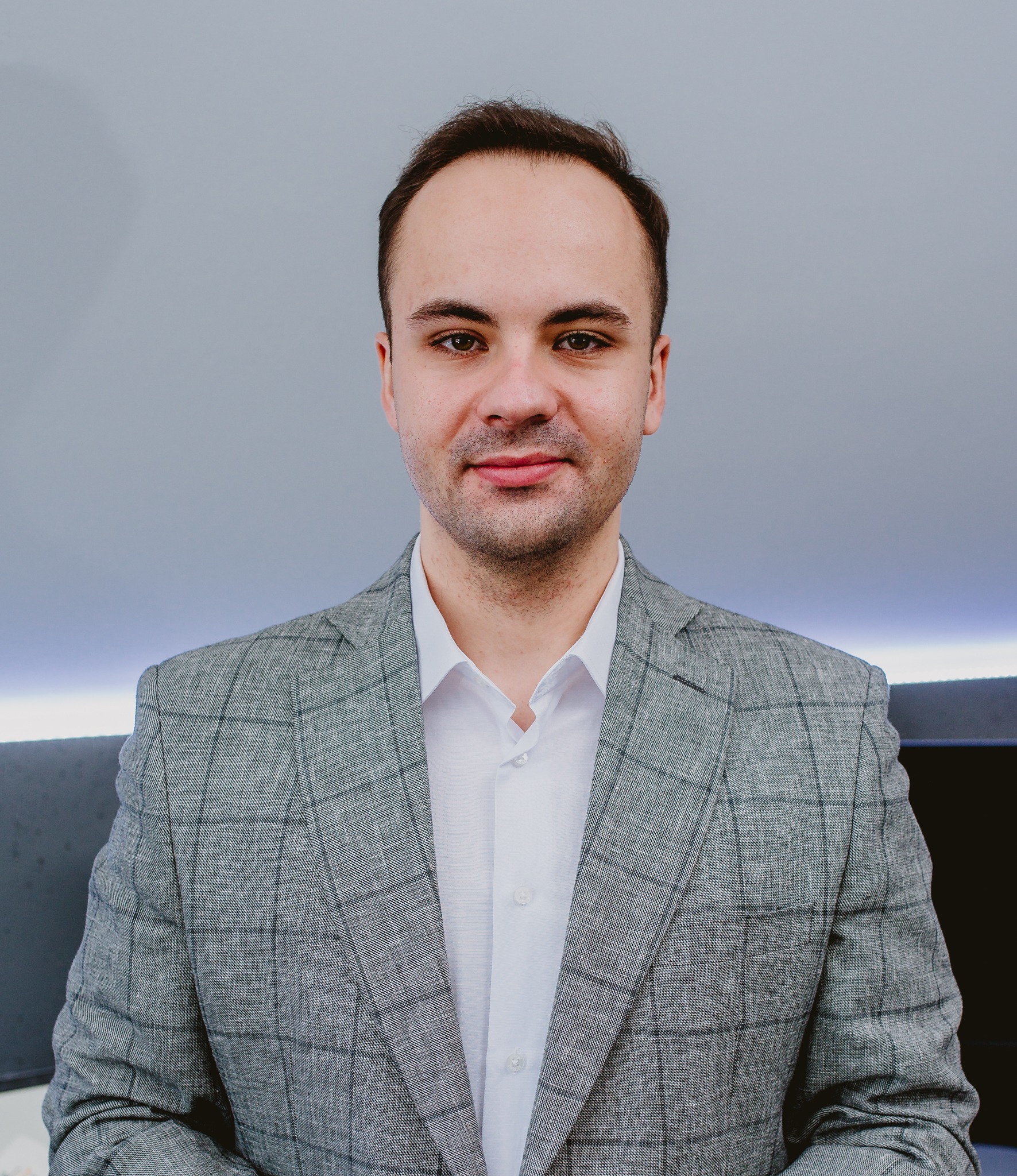 Portret mężczyzny w szarej marynarce w kratę i białej koszuli, z delikatnym uśmiechem na twarzy, na tle szarej ściany z subtelnym oświetleniem.